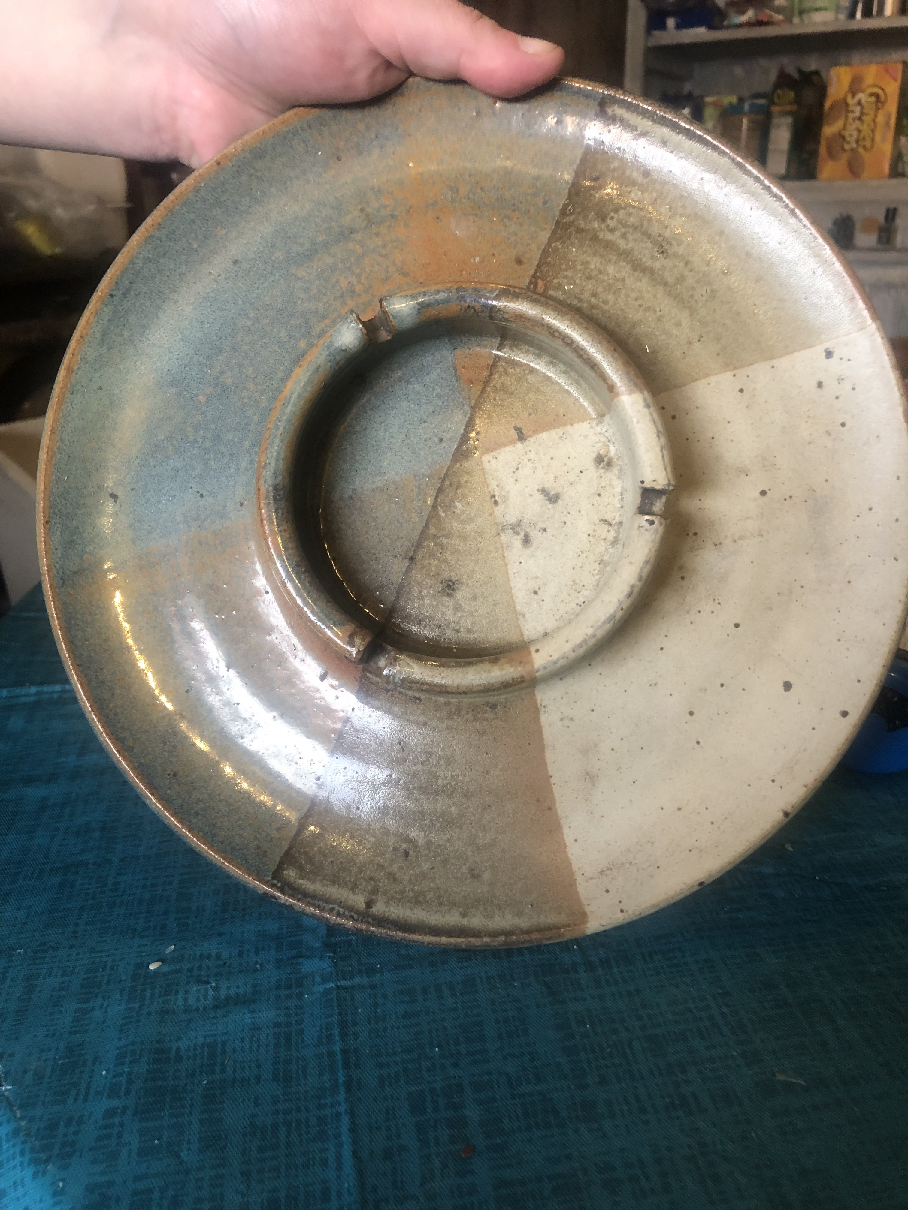 A picture of a circular ashtray being held upright, it is a big circular dish, with a raised wall with three evenly spaced notches for joints. It is glaced in oxides and glaze in blue, teal, brown and grey.